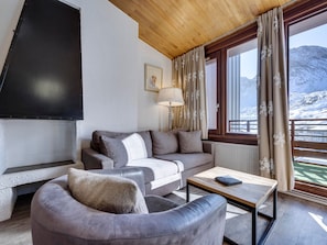 Un salon moderne comprenant un canapé, un fauteuil et une table basse, un téléviseur à écran plat fixé au mur et une fenêtre avec des rideaux.