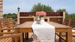 A wooden table with a white tablecloth, surrounded by wooden chairs, set against a tiled wall with a view of greenery. - Verdal, quiet location near Portopetro, private pool, air conditioning, Wi-Fi (S'Alqueria Blanca)