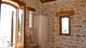 A rustic room with stone walls, a wooden ceiling, a wooden chair, a white wardrobe, a window, and a table with towels and a bowl. - Beautiful Stone Cottage with Sea and Mountain Views (Μελιδόνι)