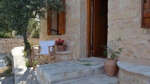 A stone building with a wooden door, a small table with a potted plant, and a chair. - Beautiful Stone Cottage with Sea and Mountain Views (Μελιδόνι)
