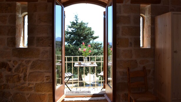 A stone-walled room with a balcony, a wooden door, and a view of trees and mountains. - Beautiful Stone Cottage with Sea and Mountain Views (Μελιδόνι)