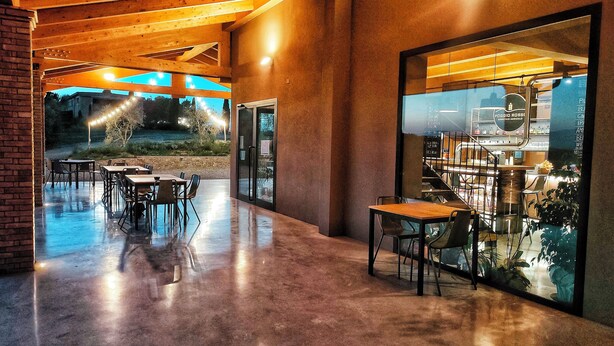 Un espacio interior con piso de concreto, vigas de techo de madera y pared de vidrio con vista a un área de bar.