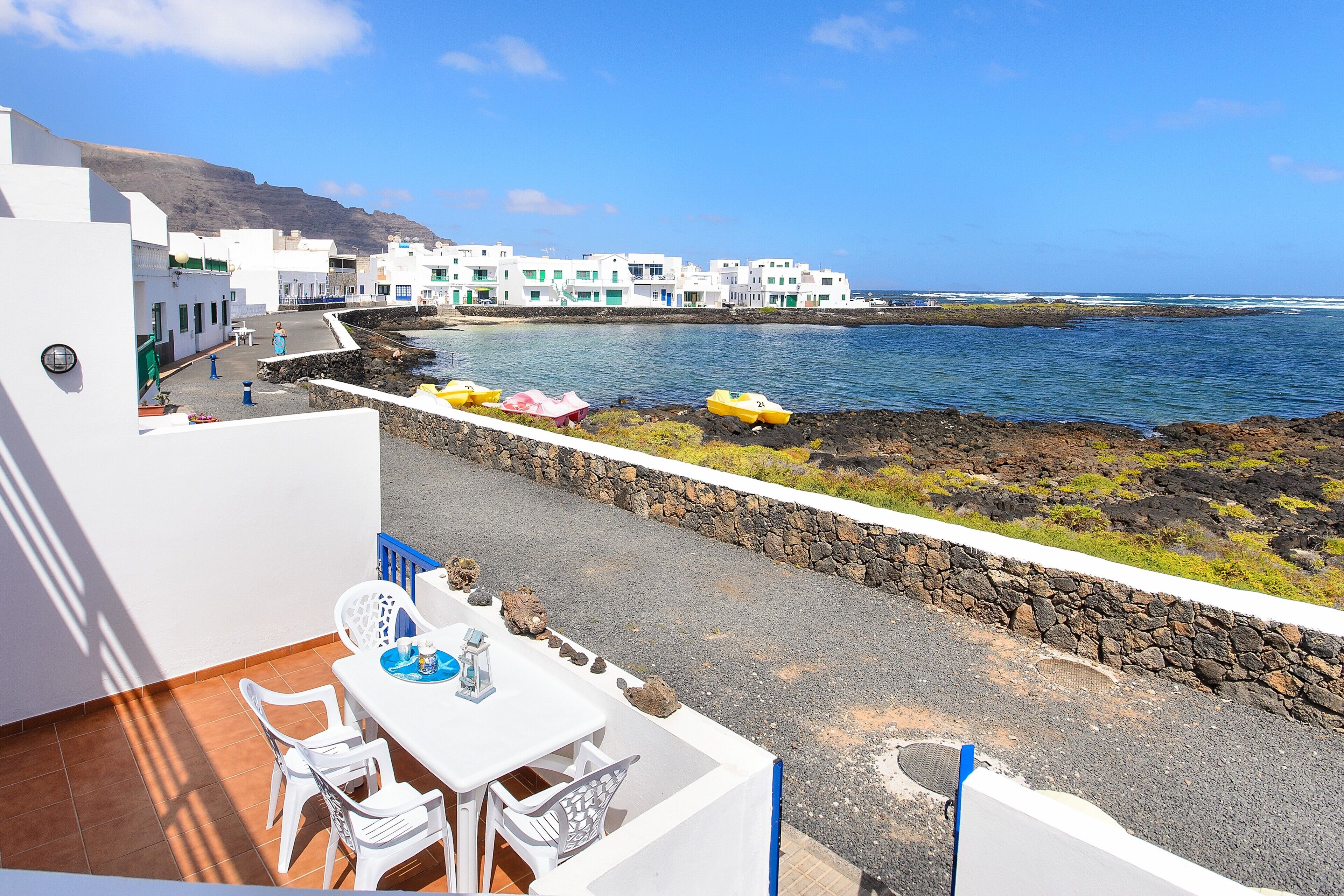 A terrace with white furniture, a view of the sea, and white buildings in the background.