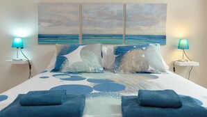 Un lit recouvert d’une couvre-lit à motifs bleus et blancs, deux lampes de chevet et un tableau représentant une scène de plage accroché au mur.