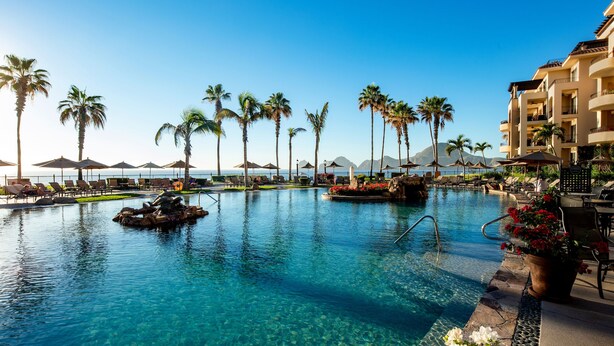 A clear blue pool surrounded by palm trees and lounge chairs, with a building in the background. - Luxury 3br villa on Medano Beach w/resort amenities! (Cabo San Lucas)
