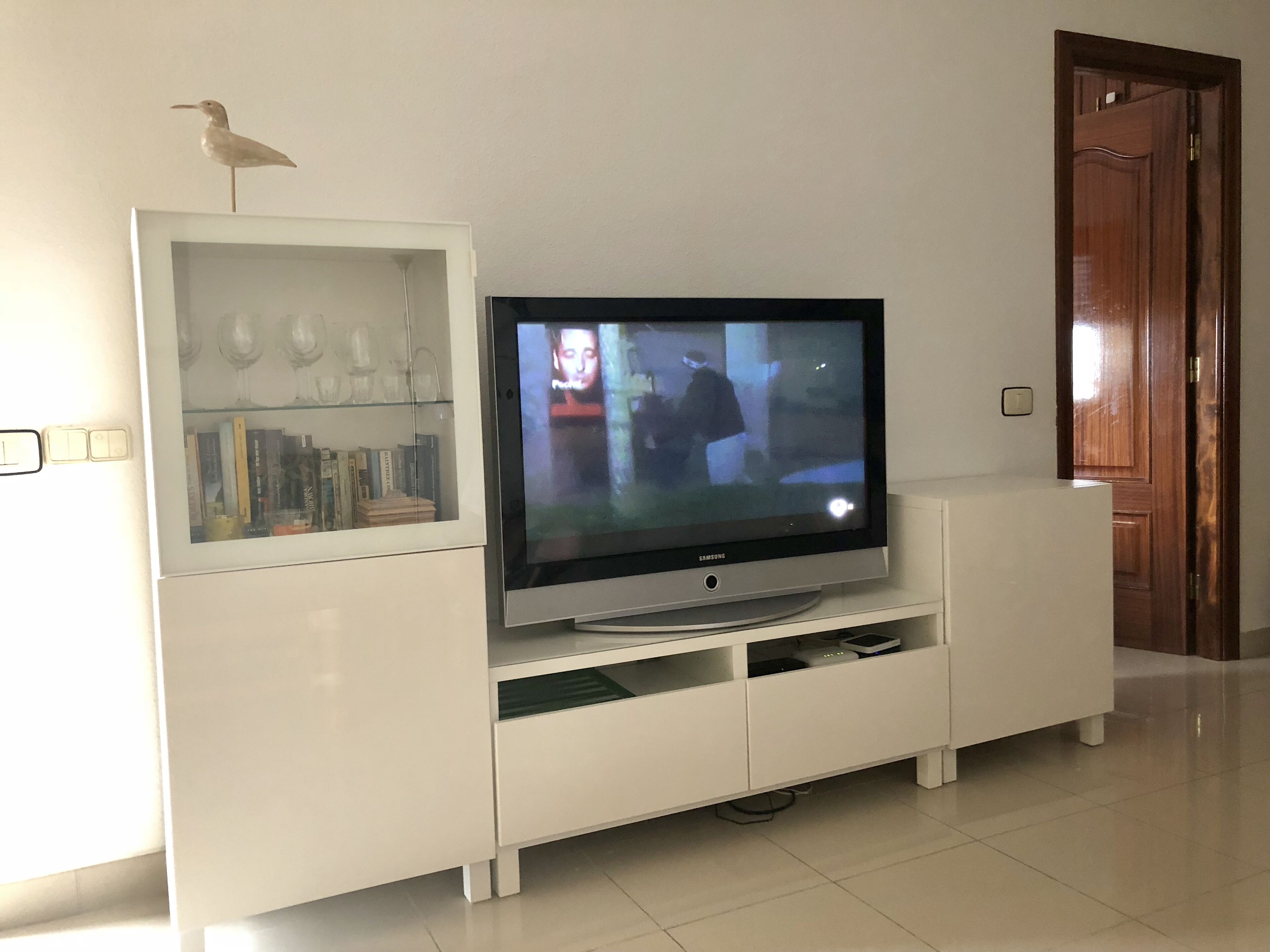 Un meuble de rangement blanc pour appareils multimédias, avec un téléviseur à écran plat, une petite étagère ornée d’objets décoratifs et une porte en bois en arrière-plan.