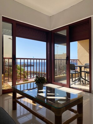 A balcony with a glass table and chairs, a potted plant, and a view of the sea.