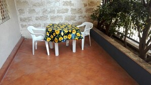 Une table ronde recouverte d’une nappe à motifs de tournesols, entourée de chaises blanches, placée devant un mur de pierre avec un sol carrelé.