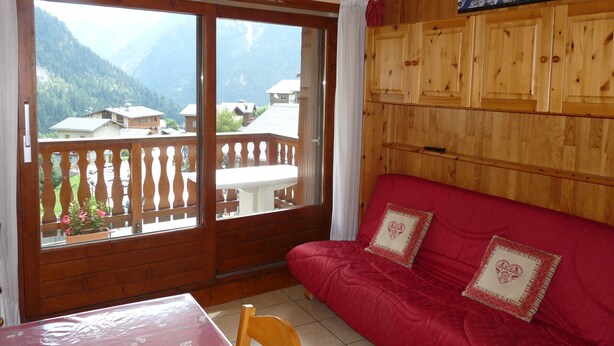 Une pièce avec un canapé rouge, des meubles en bois, une table recouverte d’une nappe à motifs floraux et une vue sur les montagnes, visible à travers de grandes portes coulissantes.