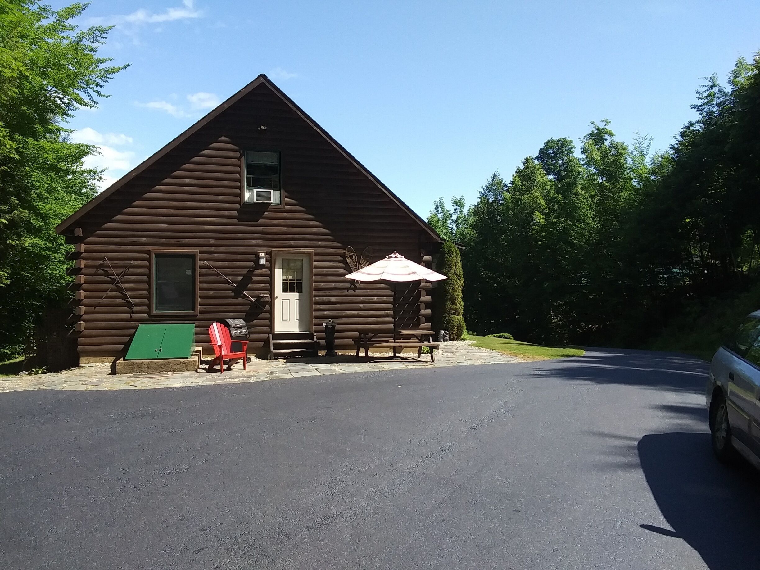 A log cabin with a porch, a red chair, a green table, and a white umbrella.