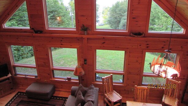 A cabin with multiple large windows offering views of a forested area. There is a ceiling fan, a lamp, and a television on a shelf.