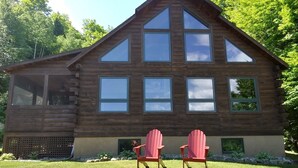 A log cabin with a gabled roof, multiple windows, and a small porch area with two Adirondack chairs.