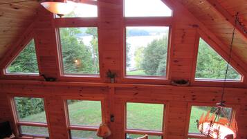 Cabana com várias janelas amplas que oferecem vista para uma área arborizada. Há um ventilador de teto, uma luminária e uma televisão em uma prateleira.