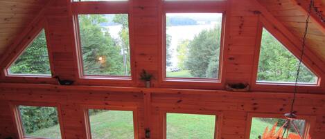 A cabin with multiple large windows offering views of a forested area. There is a ceiling fan, a lamp, and a television on a shelf.