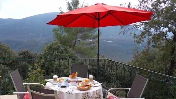 Une table ronde recouverte d’une nappe blanche, entourée de quatre chaises dotées de coussins rouges, est placée sur un balcon agrémenté d’un parasol rouge.