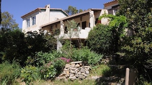 Un bâtiment à deux étages, avec un mur de pierre en premier plan, entouré d’une végétation luxuriante et d’arbres.