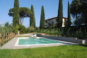 A swimming pool with a wooden deck and a stone border, surrounded by tall trees and a building in the background.