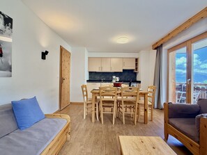 Un salon moderne avec un canapé en bois, une table à manger avec des chaises, un coin cuisine doté de meubles en bois et une grande fenêtre offrant une vue dégagée.