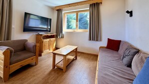 A furnished living room with a television, wooden furniture, a window with curtains, and a view of trees outside. - Pracondu 1 107 - OUTDOOR & FUN  charming apartment (Haute-Nendaz)