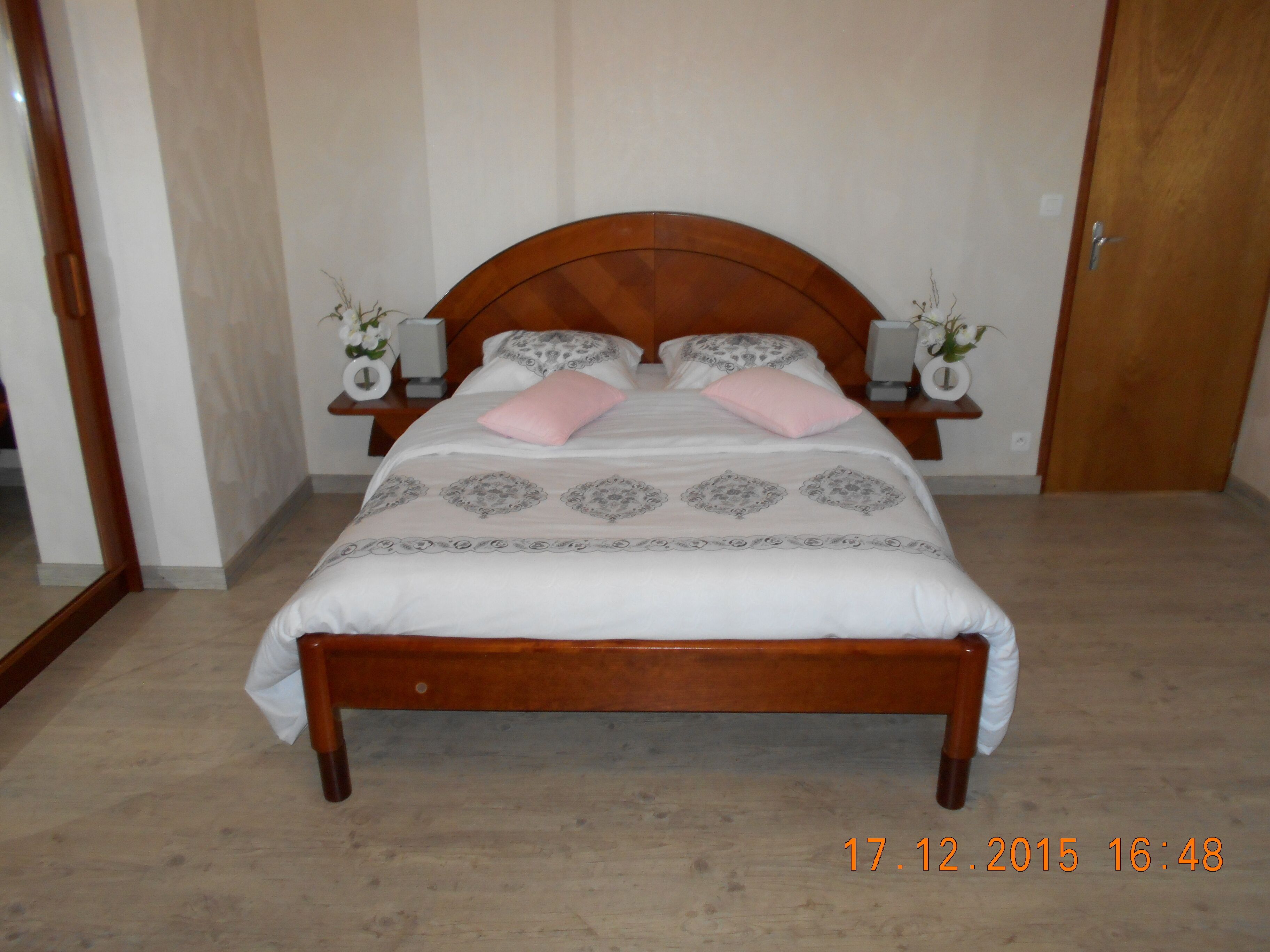 Quarto com cama de madeira, duas mesas de cabeceira com abajures e um espelho na parede esquerda.