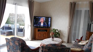 A living room with a television, sofa, chairs, a table with a vase, and a sliding glass door. - Bed & Breakfast (Villers-Sur-Port)