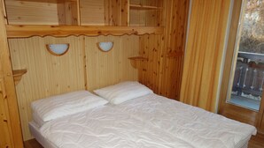 Une pièce en bois avec un lit, deux étagères fixées au mur et une fenêtre donnant sur une terrasse en bois.