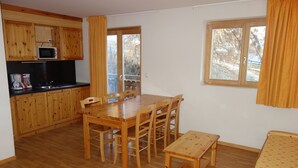 Une pièce de taille modeste comprenant une table à manger en bois, des chaises, un petit coin cuisine, un lit et une fenêtre avec des rideaux.