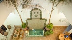 Une pièce aux hauts plafonds, dotée d’un espace salon central comprenant un canapé à motifs, une petite table agrémentée d’objets décoratifs et un élément aquatique.