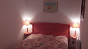 A small, cozy bedroom with a wooden headboard, a quilted bedspread, two bedside lamps, and a framed picture on the wall.