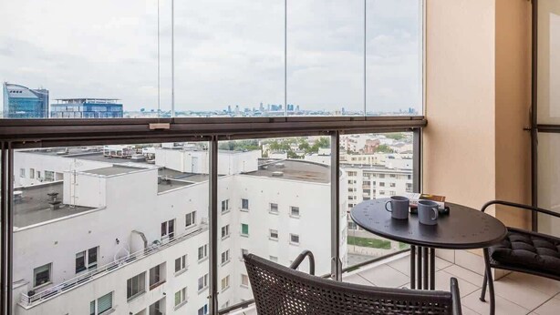 A balcony with a view of a cityscape, a round table with two chairs, and a small table with two cups.