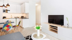 A modern kitchen with white cabinets, a dining area with colorful chairs, a TV on a stand, and a small plant on a side table.