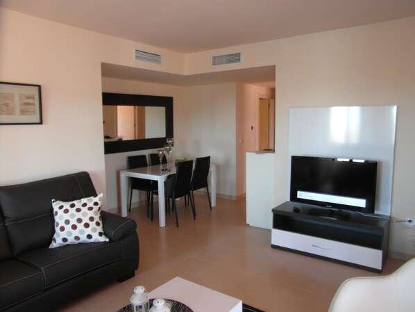 A modern living room with a black sofa, a white coffee table, a flat-screen TV, and a dining area with a white table and black chairs.