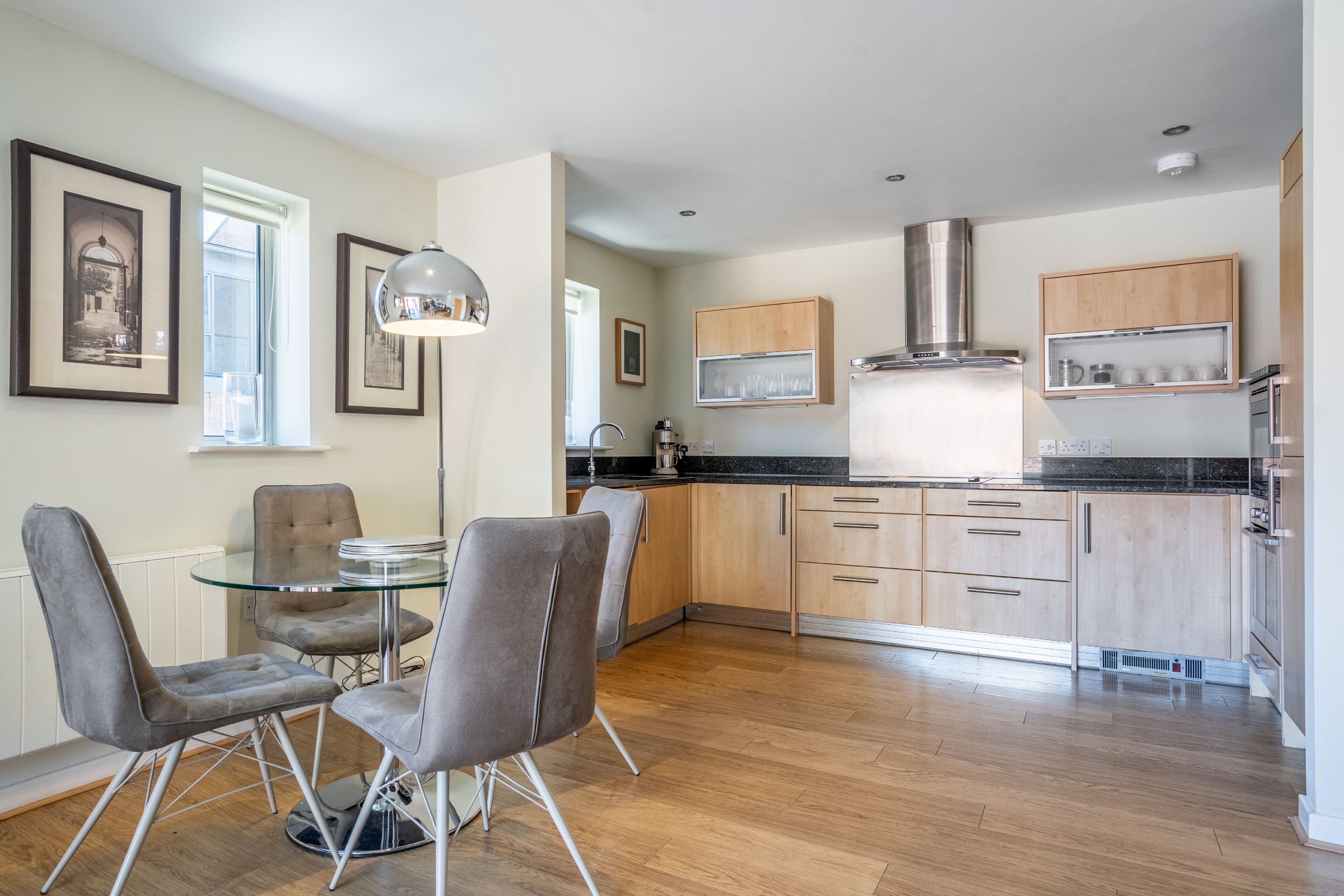 A modern kitchen with wooden cabinets, a stainless steel range hood, and a dining area with grey upholstered chairs and a glass table.
