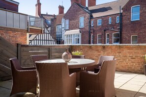 A patio with wicker furniture and a round table, brick buildings in the background, and a wooden fence.