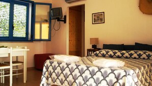 Quarto com cama, televisão na parede, janela com persianas, mesa de cabeceira e abajur.