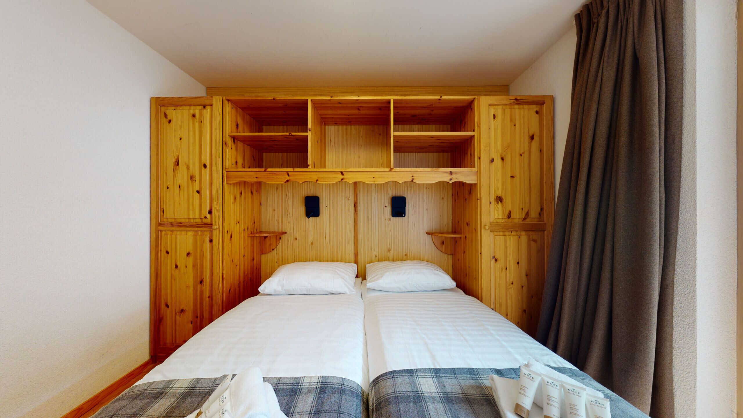 Een tweepersoonsbed met twee kussens, een houten hoofdbord met ingebouwde planken en een bruin gordijn.