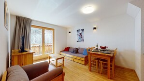 Un salon chaleureux avec un plancher en bois, un canapé, une table à manger avec des chaises, une fenêtre avec des rideaux et un luminaire fixé au mur.
