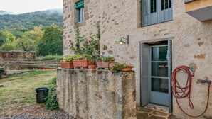 Un edificio de piedra con puerta, ventanas y una manguera. Hay plantas en macetas sobre un alféizar y un cubo negro en el suelo.