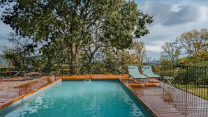 Una piscina rectangular con una cubierta de madera, rodeada por una cerca metálica y árboles.