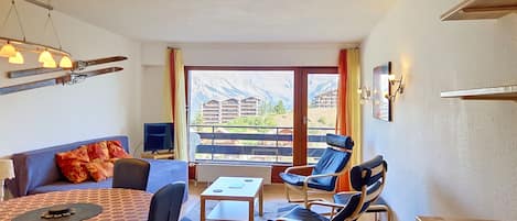 Ein Zimmer mit einer Couch, zwei Stühlen, einem kleinen Tisch, einem Fernseher und einem Balkon mit Bergblick.