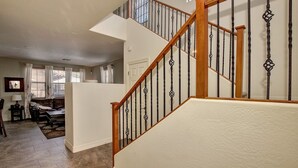 A spacious living room with a high ceiling, a staircase with wooden railings and black metal balusters, and a view of the outside through large windows.