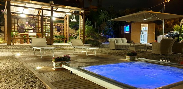 A well-lit outdoor area with a hot tub, lounge chairs, and a wooden pergola.