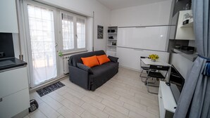 A compact living space with a sofa, a small table with flowers, a kitchenette, and a sliding door to a balcony. - FIUMICINO BUSINESS & VACATION APARTMENT CIR 5572 (Fiumicino)