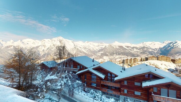 A wooden lodge with multiple balconies, set against a backdrop of snow-covered mountains and clear skies.