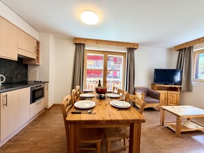 Un coin repas avec une table en bois dressée pour quatre personnes, une cuisine équipée d’une cuisinière et de placards, un téléviseur posé sur un meuble et une fenêtre avec des rideaux.
