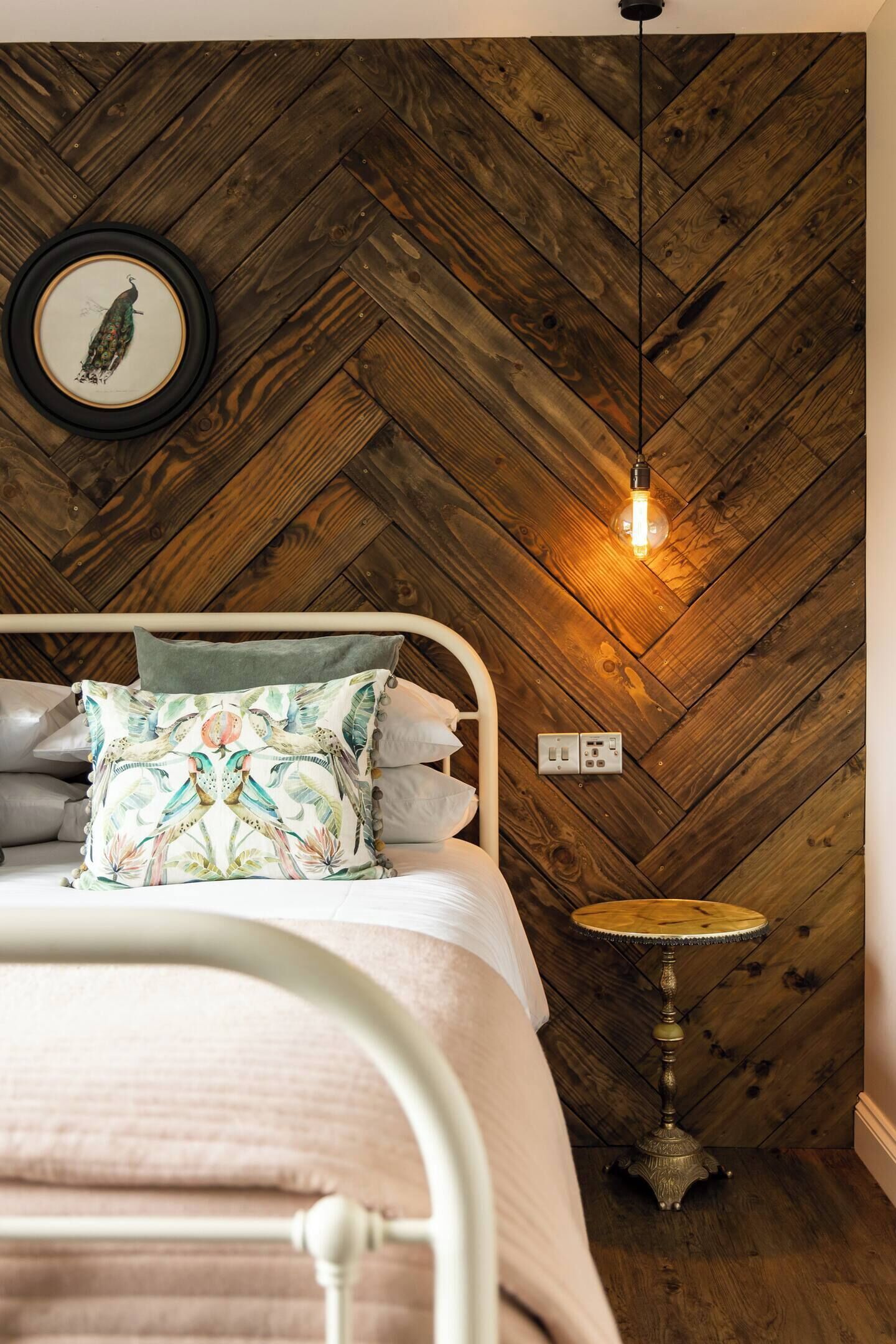 A bedroom with a herringbone wooden ceiling, a bed with a floral pillow, a bedside table, and a hanging light fixture.