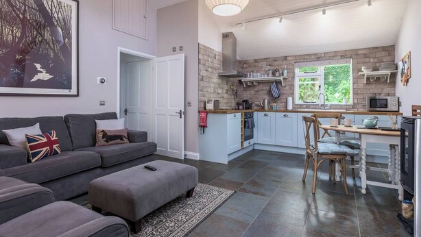 A modern living room with a grey sofa, ottoman, and a large painting on the wall. A kitchen with light blue cabinets and a dining area with a wooden table and chairs is visible. - Pengelly is Laid back luxury in 9-acres of countryside right near the beach (Kilkhampton, Bude)