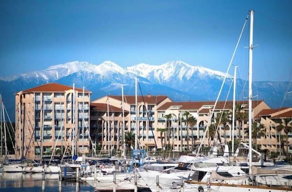 Un port de plaisance où sont amarrés de nombreux bateaux, une rangée d’immeubles dotés de balcons et, au loin, une chaîne de montagnes.