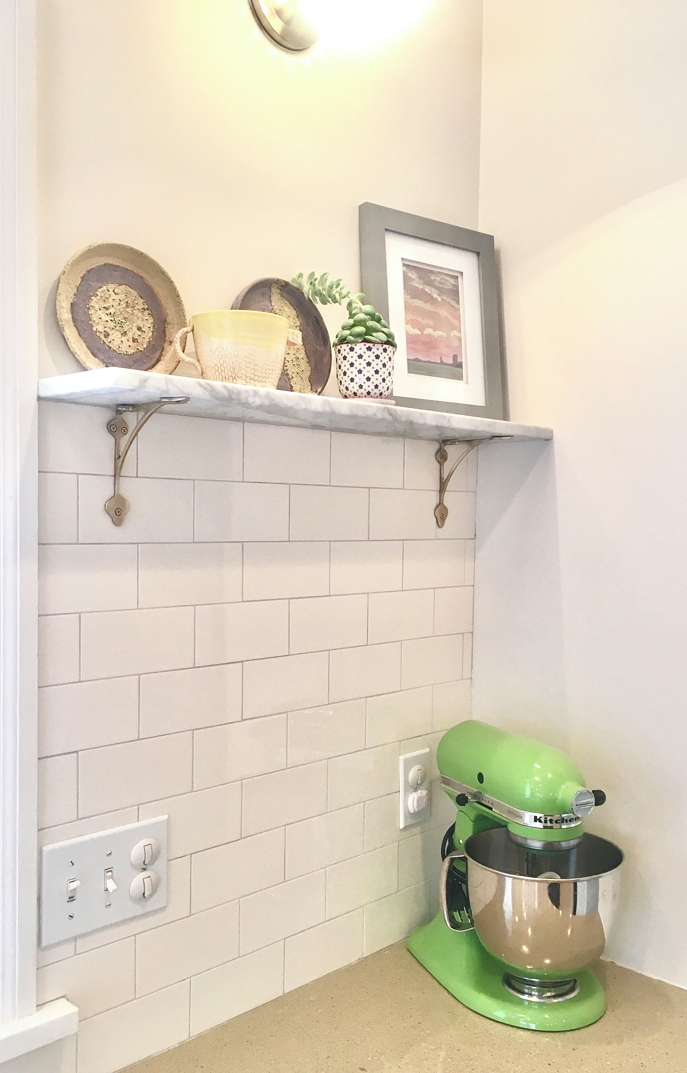 A kitchen with white subway tiles, a green stand mixer, and a shelf with decorative items.