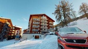 A multi-story wooden building with balconies, a snow-covered slope, and a red car parked in front. - Pracondu 1 306 - OUTDOOR & FUN  charming apartment (Haute-Nendaz)